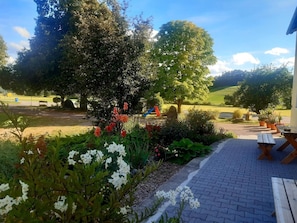 A paved outdoor area with a picnic table and benches, surrounded by lush greenery and flowering plants.