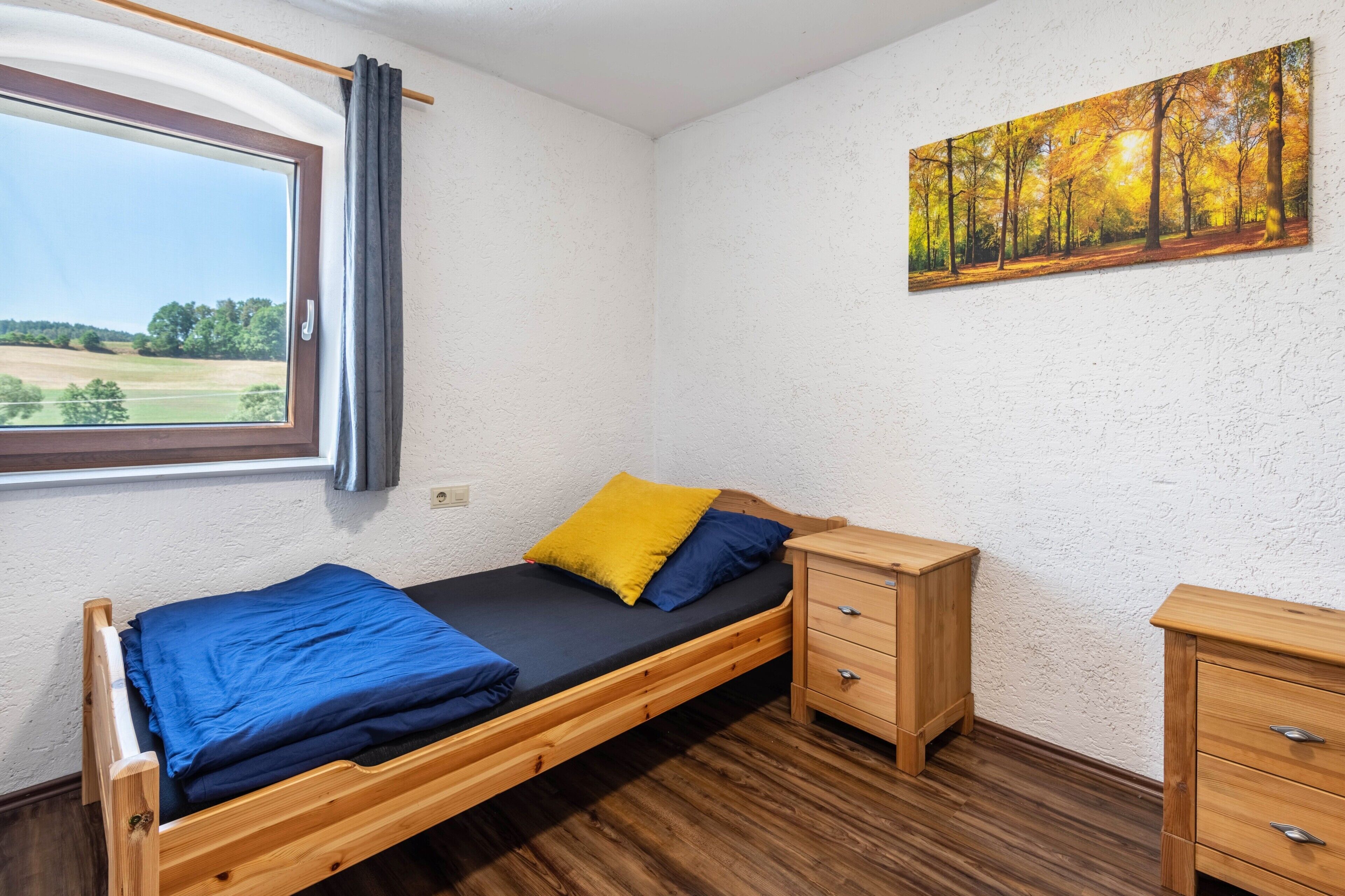 Ein Einzelbett mit Holzbettrahmen, ein Nachttisch, eine kleine Kommode und ein Fenster mit Blick auf ein Feld.