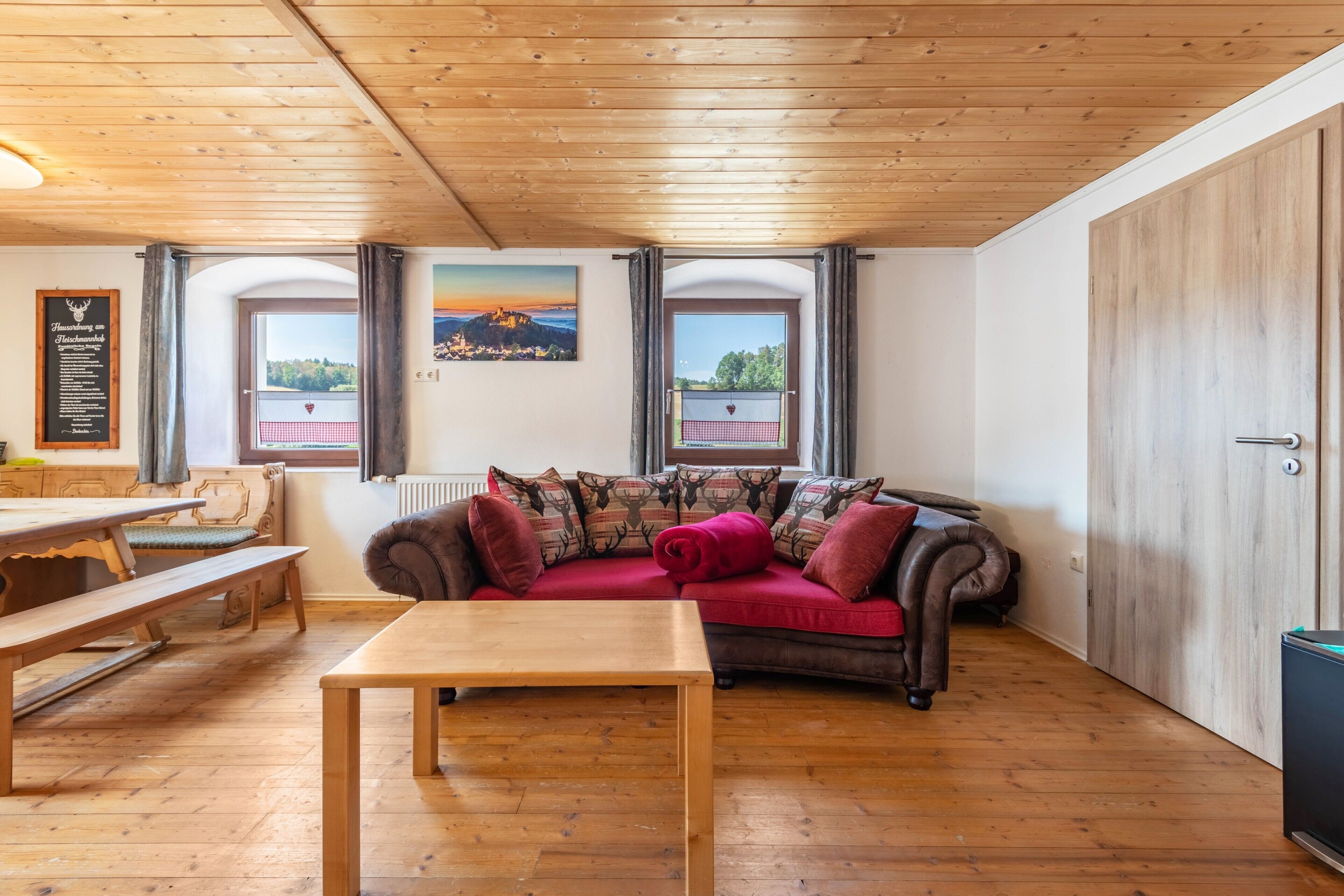 A cozy room with a wooden ceiling, a sofa with patterned cushions, a wooden table, and a view through a window.