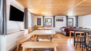 A cozy room with a wooden ceiling, a sofa, a dining table with benches, and a TV mounted on the wall.