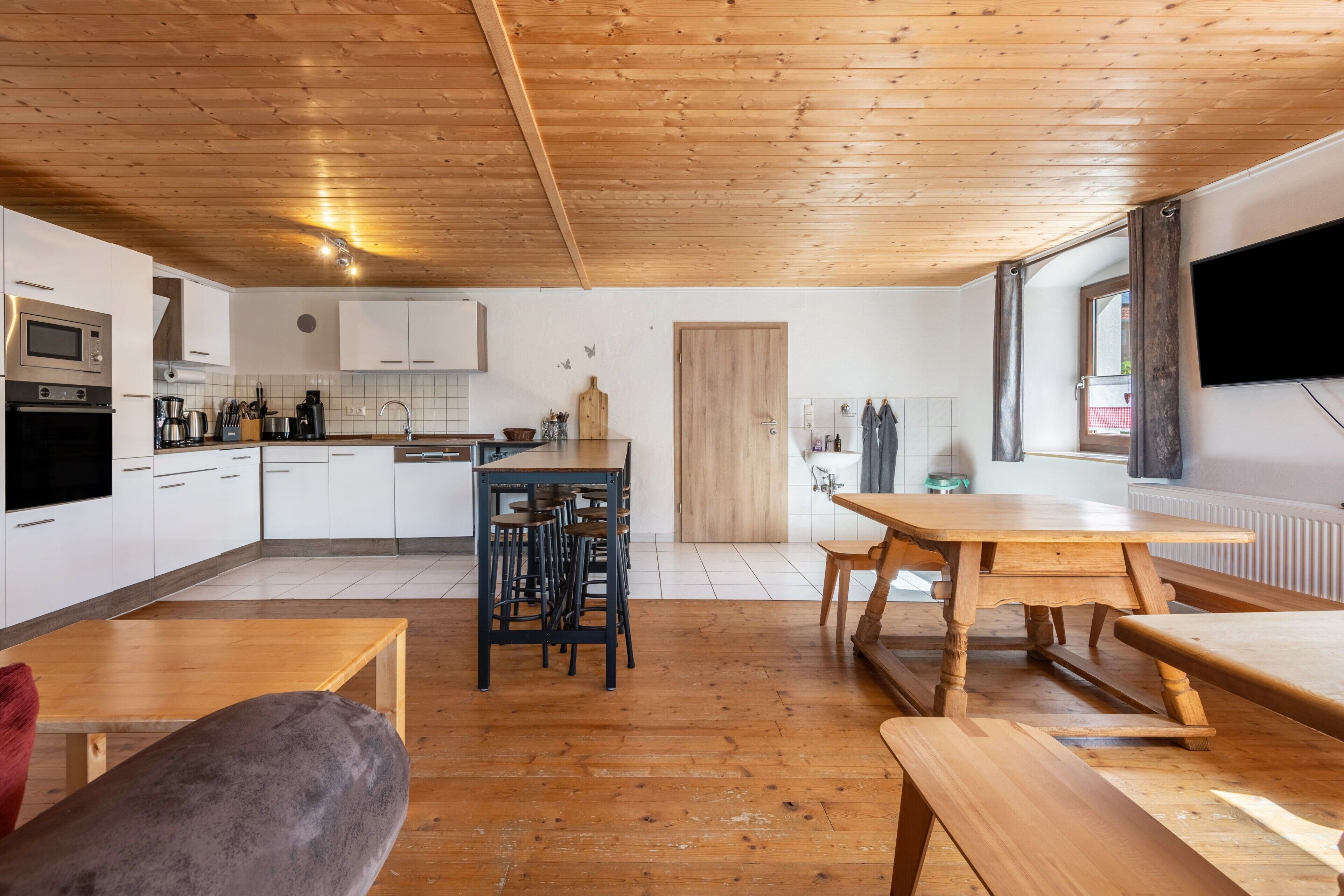 A modern kitchen with white cabinets, a wooden ceiling, and a dining area with a wooden table and benches.