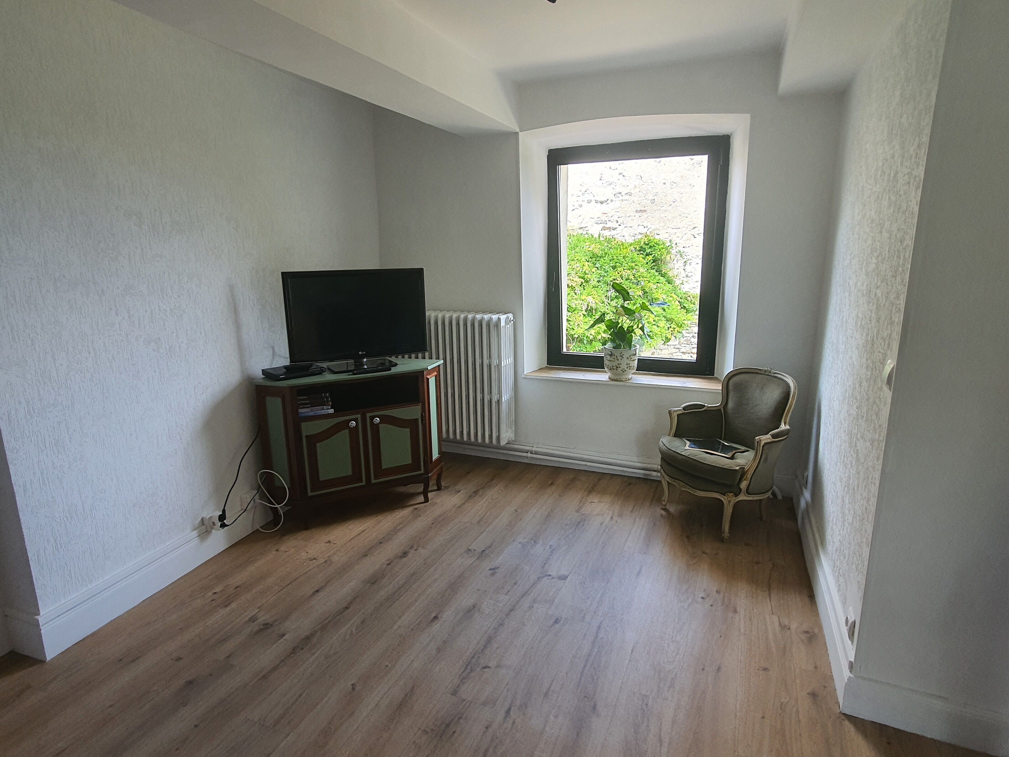 Une pièce avec un tél éviseur posé sur un meuble en bois, un radiateur, une fenêtre donnant sur un espace verdoyant et une chaise.