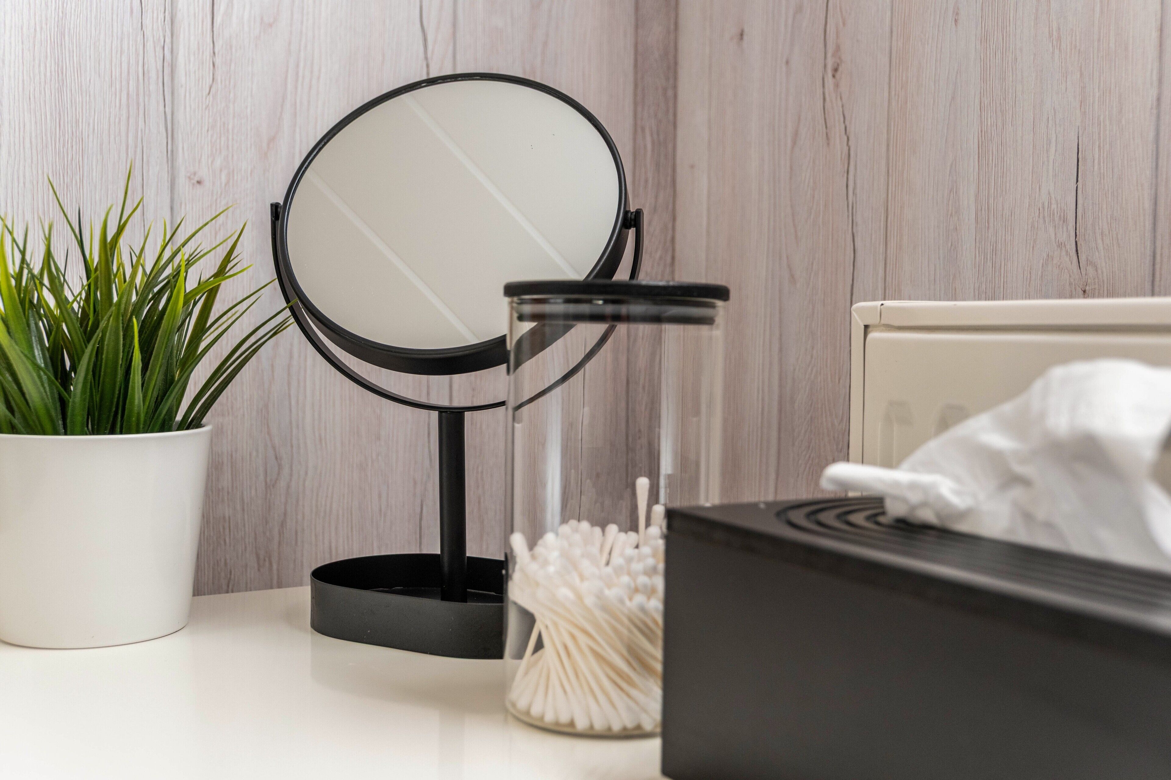 A mirror on a stand, a potted plant, and a container with cotton swabs on a table.