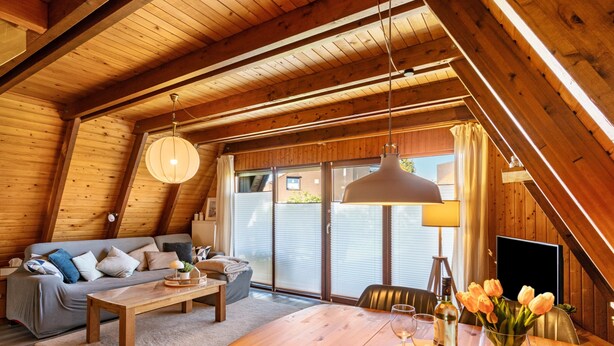 A cozy loft-style room with a wooden ceiling, a sofa, a dining table, and a large window. - Holiday Home Alma – Private Terrace, Wi-Fi, Garden & Coffee Delight Included (Wittmund)
