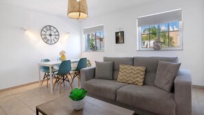 Een woonkamer met een bank, een eettafel met stoelen, een wandklok en een raam met uitzicht.