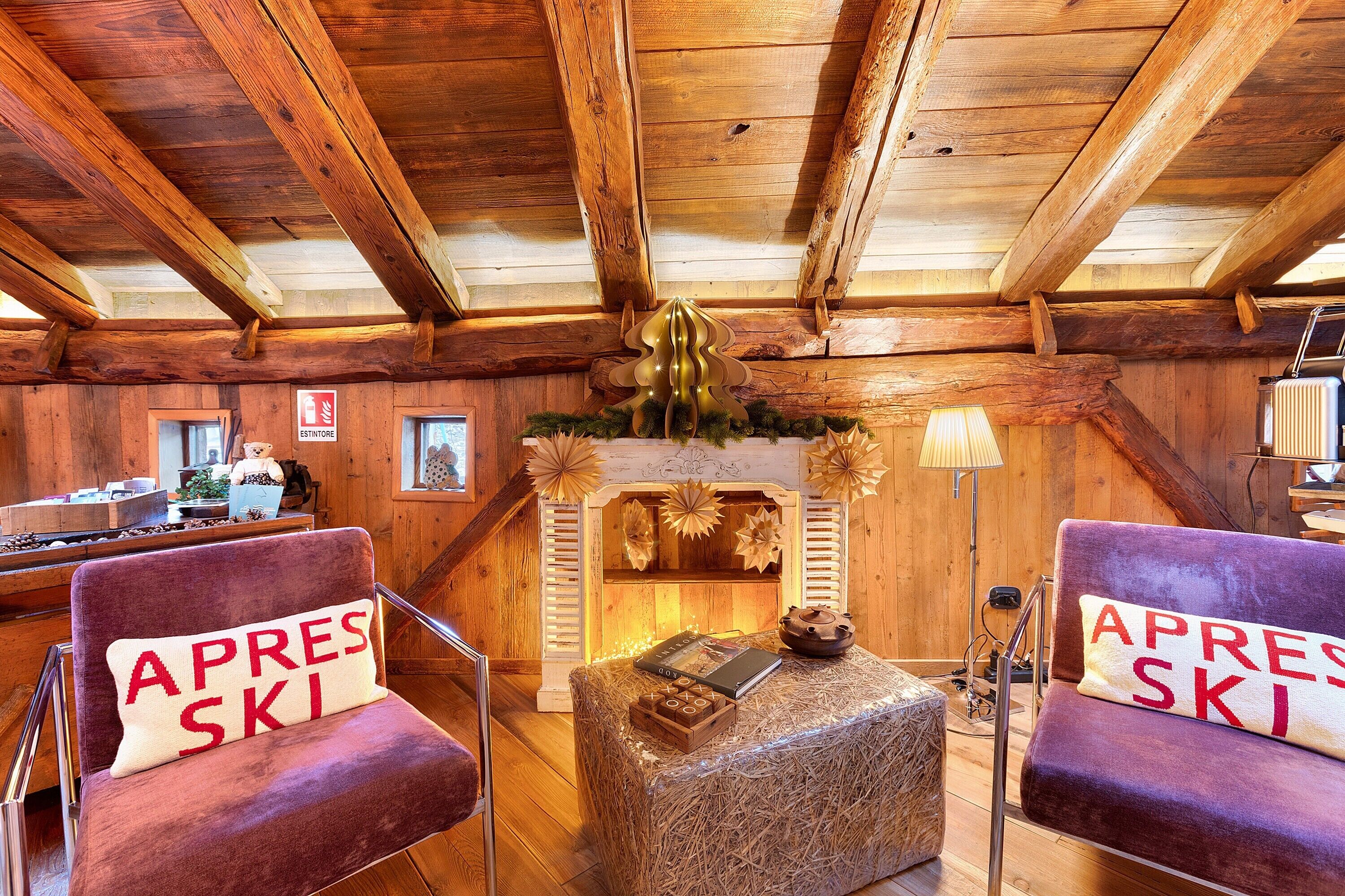 A cozy log cabin interior with wooden beams, a fireplace, and two chairs with 'APRES SKI' pillows.