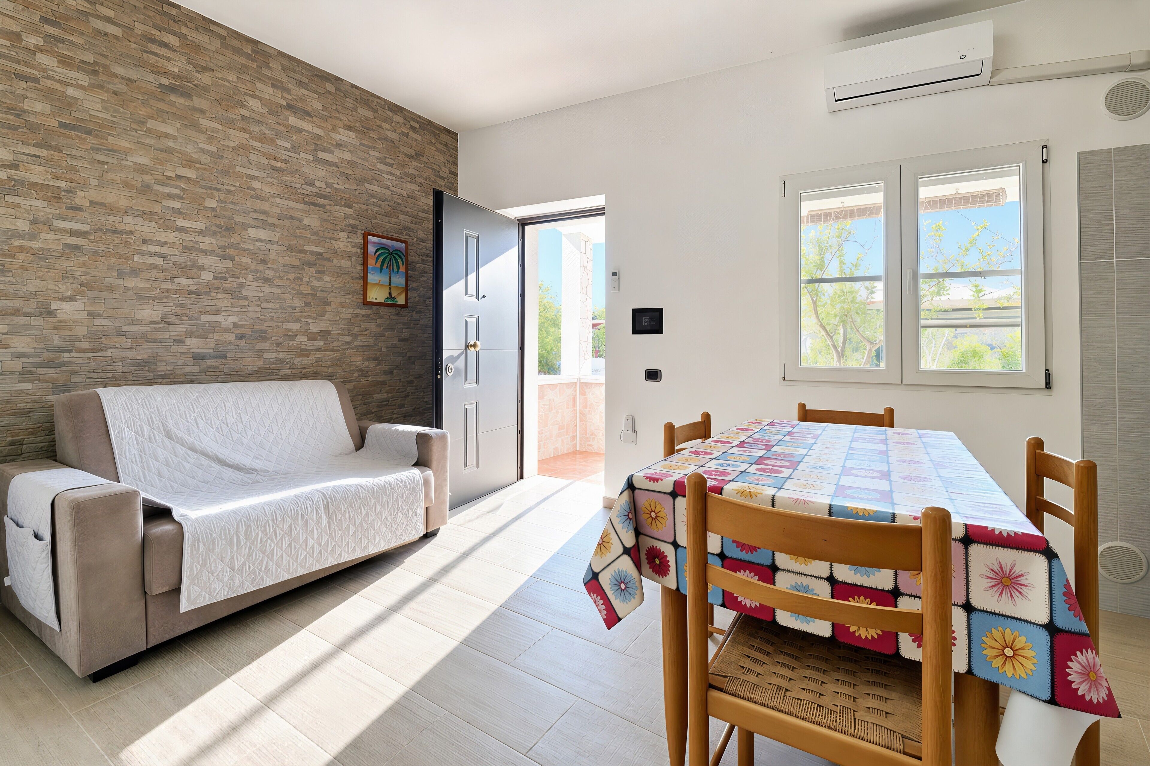 Una sala de estar moderna con un sofá, una mesa de comedor con mantel colorido, una pared de ladrillo y una puerta que da al exterior.