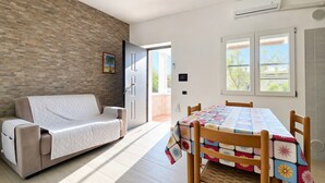 Un salon moderne avec un canapé, une table à manger recouverte d’une nappe colorée, un mur en briques et une porte donnant sur l’extérieur.