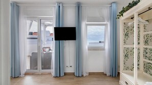 Une pièce avec un téléviseur fixé au mur, une porte-fenêtre coulissante donnant sur un espace extérieur et une armoire blanche ornée d’un papier peint à motifs floraux.