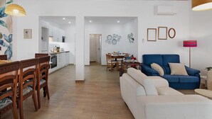 Un grand espace de vie comprenant un canapé, une table à manger et des chaises, une cuisine à l’arrière-plan et un couloir menant à une autre pièce.