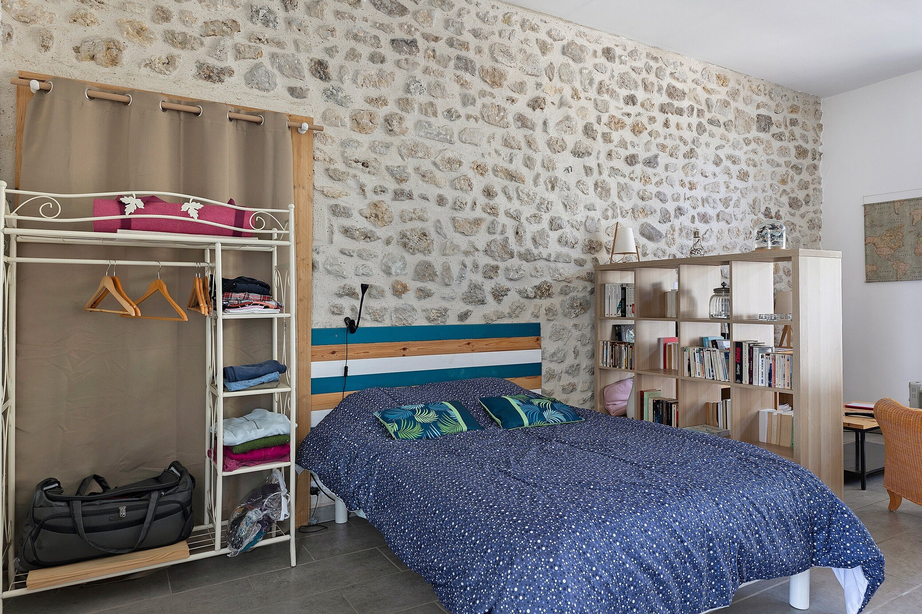 A bedroom with a stone wall, a bed with a blue patterned bedspread, a wooden wardrobe, and a bookshelf.