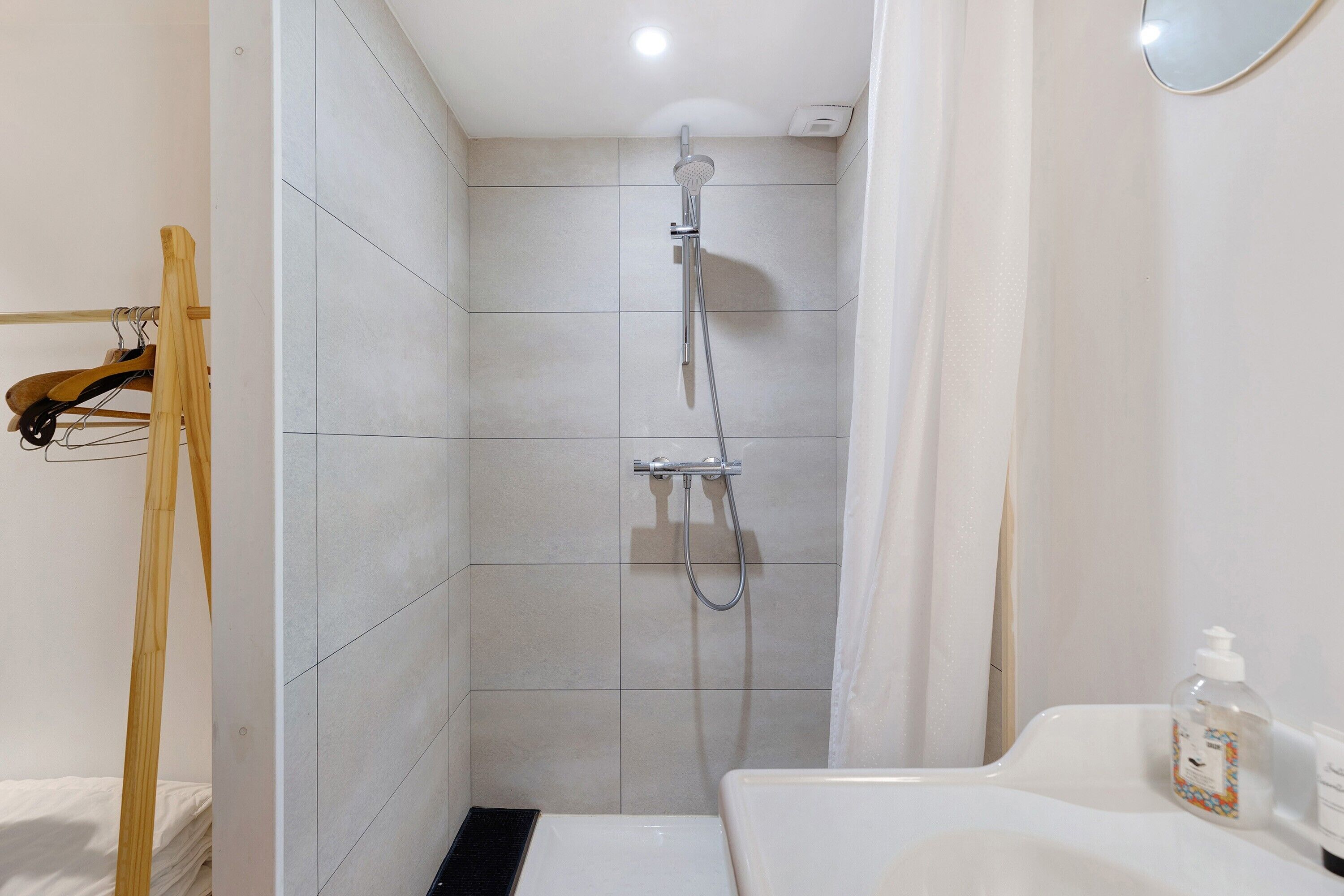 A shower with a handheld showerhead, a white sink with a soap dispenser, and a wooden rack.
