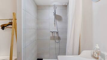 A shower with a handheld showerhead, a white sink with a soap dispenser, and a wooden rack.