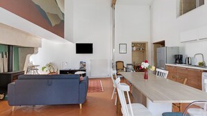 A modern living space with a sofa, dining table, chairs, a kitchen area, and a mounted TV. - Maison Bellecour: Shared Terrace, Garden & Wi-Fi in Vaudelnay (Vaudelnay)