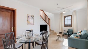 Un'area pranzo con tavolo in vetro, sedie e televisore. Si vede una scala, un divano e una finestra con tende.
