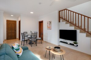 A living room with a sofa, a dining table with chairs, a TV mounted on the wall, and a staircase.