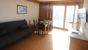 Una sala de estar con un sofá oscuro, una mesa de comedor de madera con sillas, un cuadro enmarcado en la pared y un televisor sobre un mueble de madera.