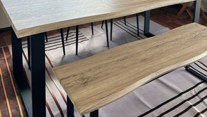 Una habitación con un ventanal grande, una mesa, sillas, un banco, una planta en maceta y un cuadro en la pared.