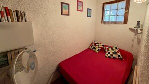 A small, cozy room with a red bed, two pillows, a window with lace curtains, and a ceiling fan. - Bright T2 Apartment - 200m from Beach, 4 pax (Argelès-sur-Mer)