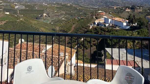 En balkon med udsigt over et bjerglandskab, en hvid stol og en bygning med rødt tag.