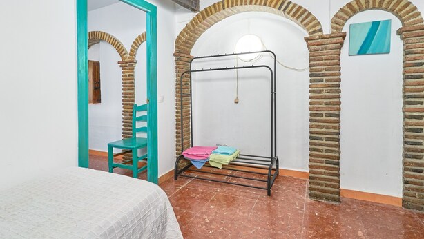 Ein Zimmer mit einem Bett, einem Sessel, einem Metallregal mit gefalteter Kleidung und Backsteinbögen.