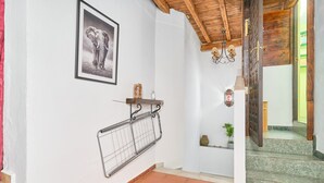 Ein Flur mit gefliestem Boden, einem gerahmten Bild an der Wand, einem Metallgestell, einem kleinen Regal mit einer Vase und einer Treppe mit hölzernem Geländer.