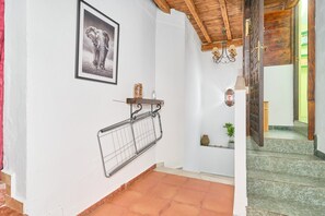 Een gang met betegelde vloer, een ingelijst beeld aan de muur, een metalen rek, een klein schap met een vaas en een trap met houten leuning.