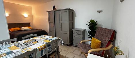 Une petite pièce bien éclairée, avec un lit, une table recouverte d’une nappe à motifs, une chaise, un meuble et une plante en pot.