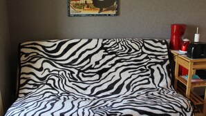 Ett rum med ett sängöverkast i zebrastripe, en affisch på väggen och ett litet träbord med en röd vas och ett svart föremål.