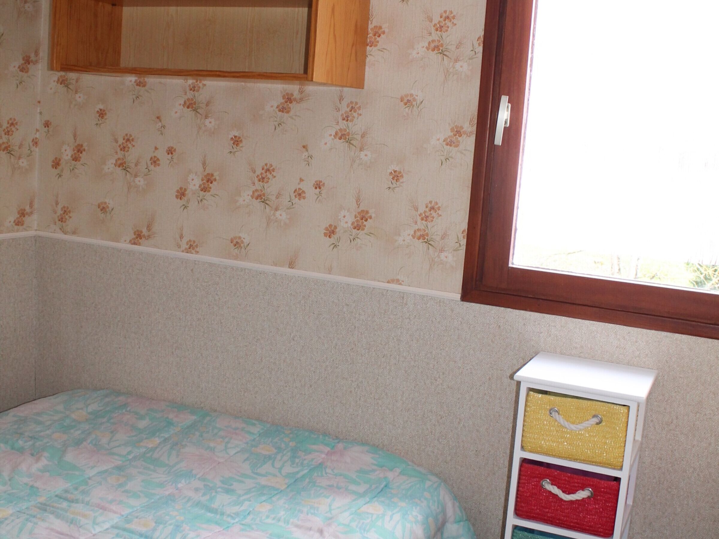 A bedroom with floral wallpaper, a bed with a patterned bedspread, a window with a view, and a small white storage unit with drawers.