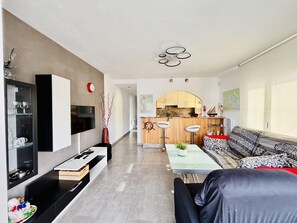 A modern living room with a sofa, a coffee table, and a TV. There is a kitchen in the background with a bar counter and stools. - Renovated Apartment in Santa Margarita - 2 Bedrooms (Roses)