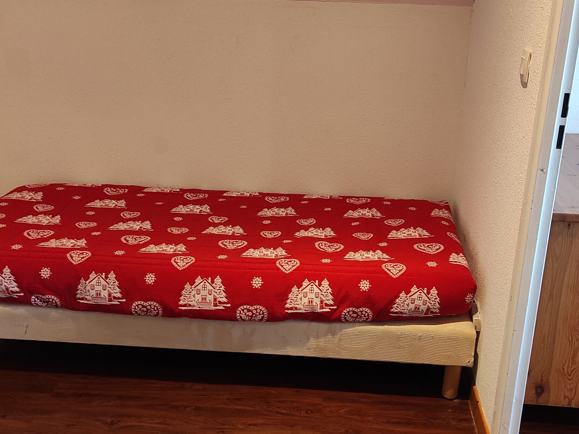 Un petit lit recouvert d’une couverture rouge ornée de motifs blancs représentant des maisons et des arbres.