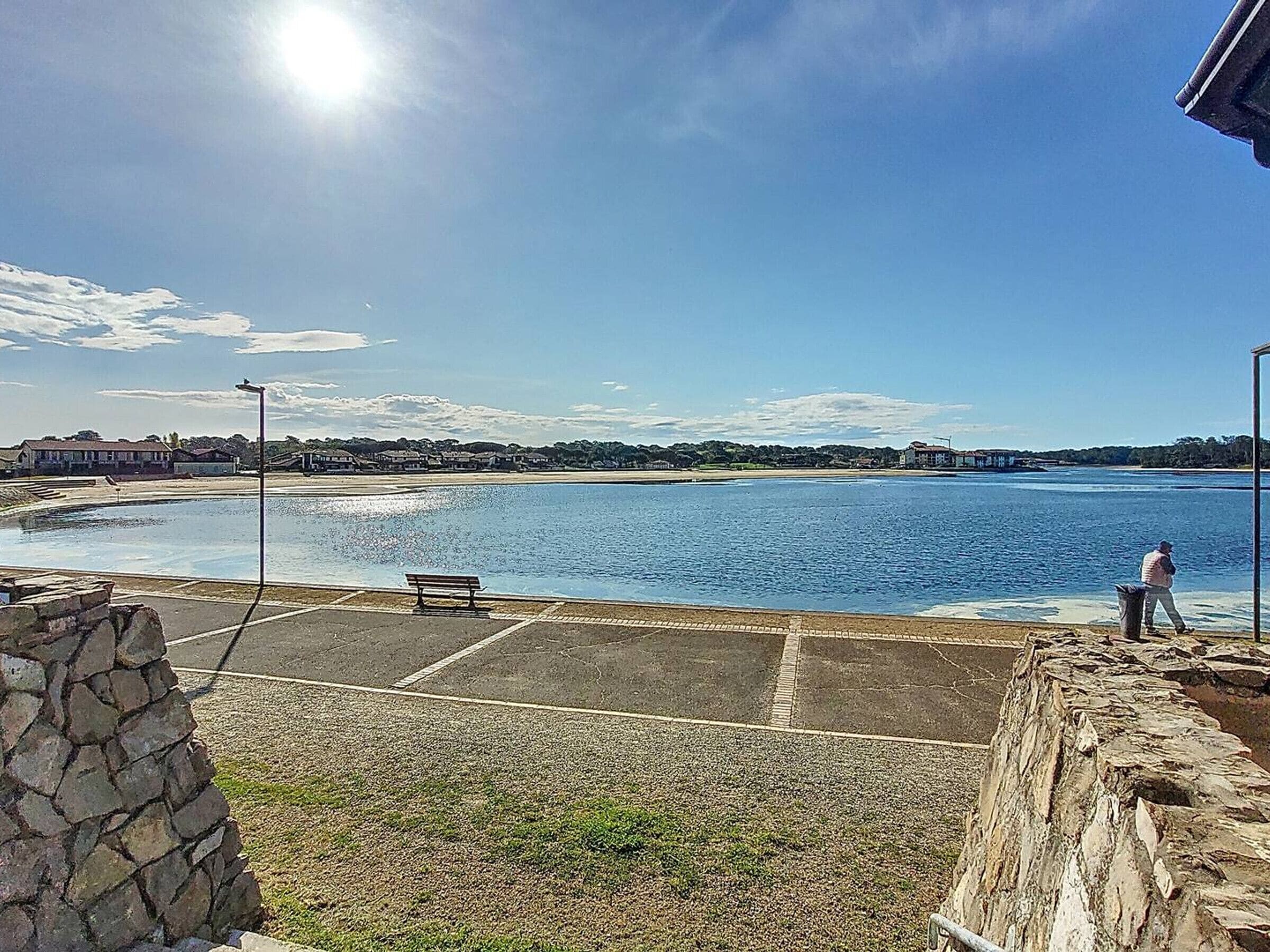 A coastal view with a bench, a stone wall, and a person walking by the water.