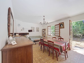 A dining area with a wooden table, chairs, and a chandelier. There is a sofa and a bookshelf in the background.
