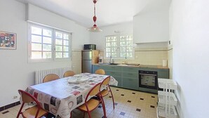A dining area with a table and chairs, a kitchen with a refrigerator and stove, and windows with a view of greenery. - Villa for 6 guests in Moliets with pets allowed (Moliets-et-Maa)