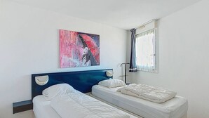 Quarto de hotel com duas camas de solteiro, cabeceira azul, pintura, janela com cortinas e mesa de cabeceira.