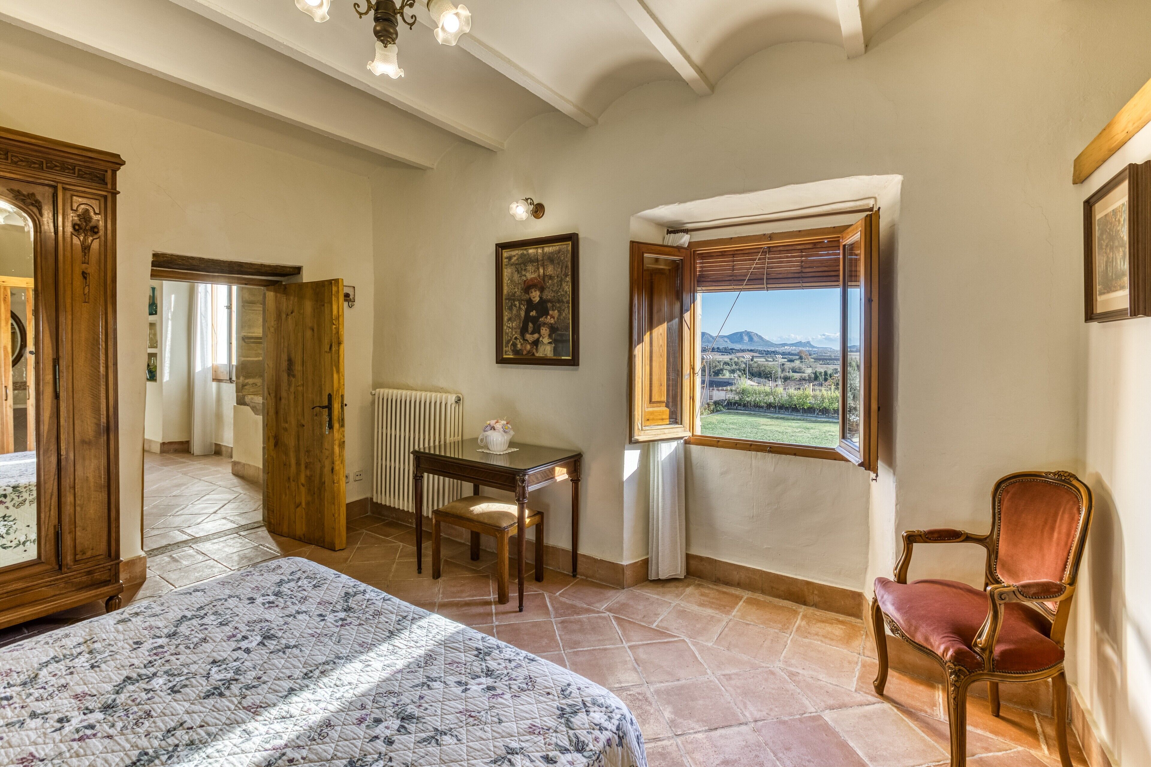 Ein Schlafzimmer mit Bett, Holzstuhl, Tisch, Heizkörper, Fenster mit Blick und einem Bild an der Wand.