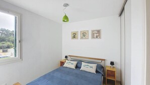 Une chambre avec un lit simple, une fenêtre, une lampe suspendue verte et trois tableaux encadrés accrochés au mur.