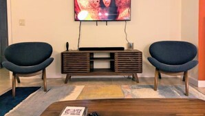 Un salon doté d’un téléviseur à écran plat fixé au mur, d’un meuble de rangement en bois en dessous, de deux fauteuils sombres et d’une table basse sur laquelle repose un magazine.