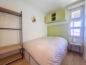 Ein Einzelbett mit Holzrahmen, ein kleiner Nachttisch, ein grünes Stauraumelement und ein Fenster mit Jalousien.
