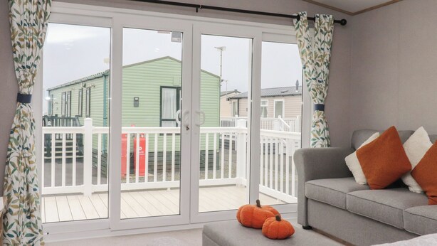 A modern living room with a grey sofa, orange cushions, and a white ottoman. There are two small orange pumpkins on the ottoman. The room has a large sliding glass door leading to a balcony with a white railing and a green building in the background. - The Retreat (Morecambe)