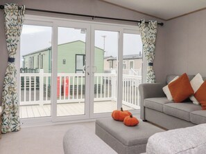 A modern living room with a grey sofa, orange cushions, and a white ottoman. There are two small orange pumpkins on the ottoman. The room has a large sliding glass door leading to a balcony with a white railing and a green building in the background. - The Retreat (Morecambe)
