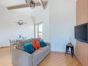 A modern living room with a grey sofa, a dining table, and a TV mounted on a wooden wall. - 2-Bedroom Apartment for 6 Guests with Pool (Vieux-Boucau-les-Bains)