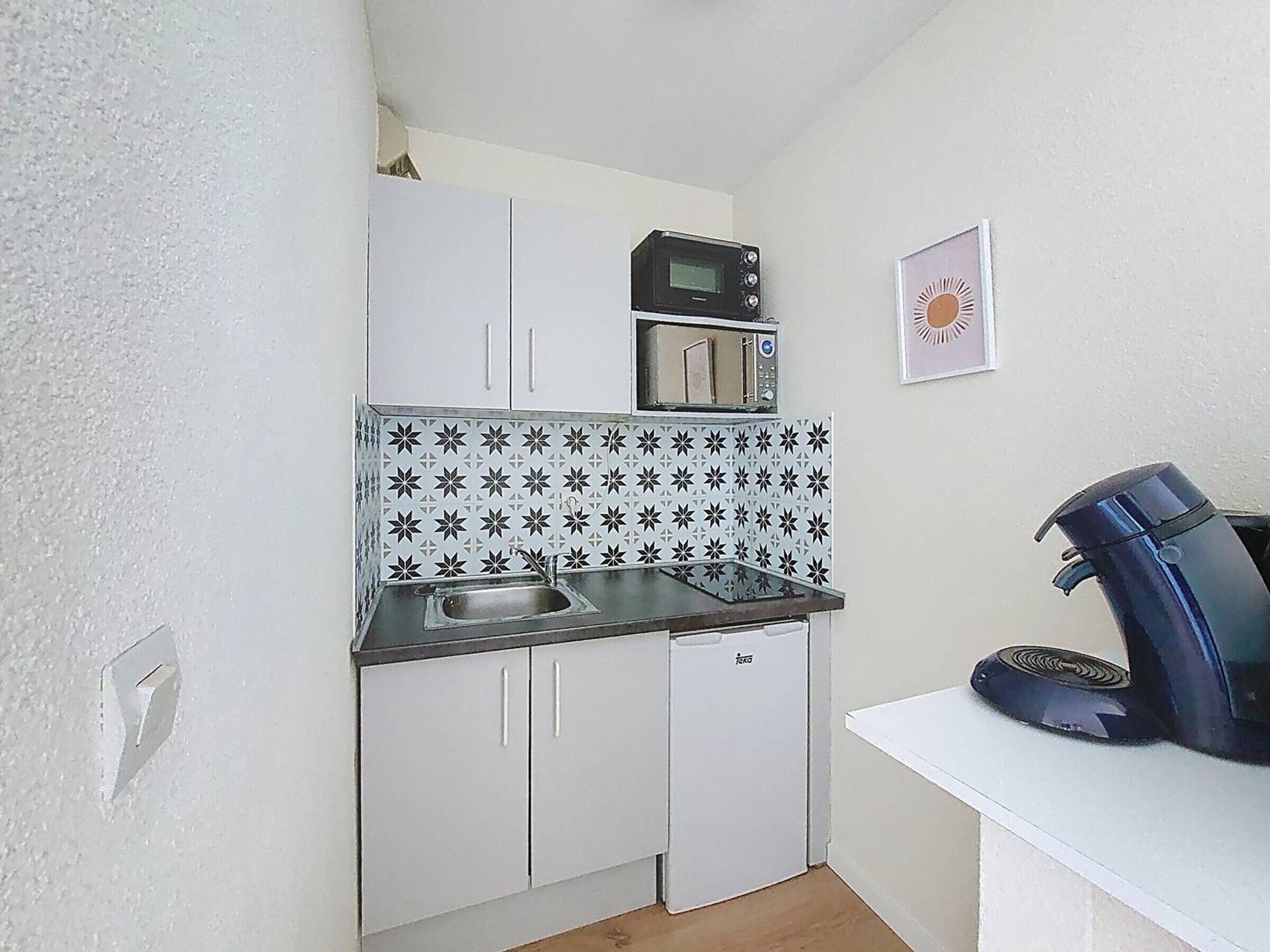 A compact kitchenette with white cabinets, a microwave, a sink, and a coffee machine.