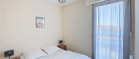 A bedroom with a bed, a nightstand, a wall-mounted lamp, a picture, and a window with curtains.