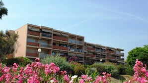 A multi-story building with balconies and a garden with pink flowers in the foreground.