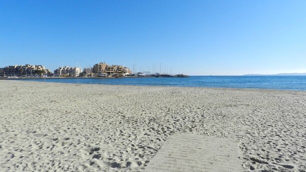 A sandy beach with a clear blue sky. In the background, there are buildings with multiple floors and balconies.
