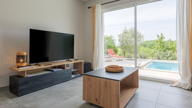 A modern living room with a flat-screen TV, wooden coffee table, and a large window overlooking a pool and greenery. - Villa Flora with Private Pool, Terrace, and Wi-Fi (Balazuc)