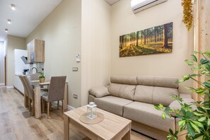 Moderni olohuone, jossa on sohva, ruokapöytä ja tuoleja. Taustalla on keittiöalue, jossa on kaappeja ja mikroaaltouuni. Seinällä on taulu, jossa on kuvattuna metsä.