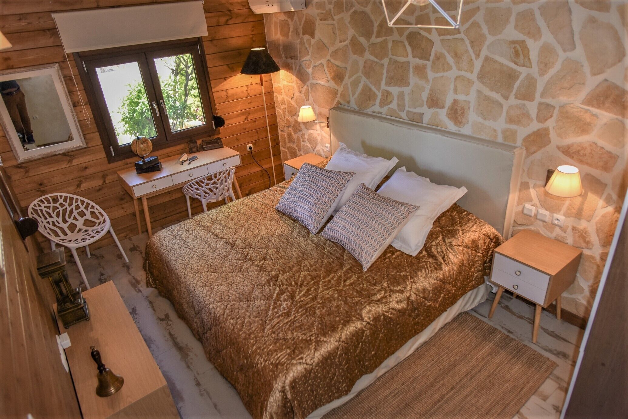 A cozy bedroom with a stone wall, wooden furniture, a bed with patterned bedding, and a desk with a chair.